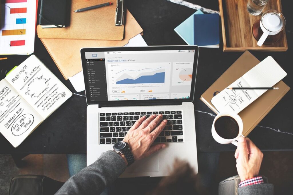 laptop with charts and graphs on a desk filled with notes and files with a man's hands holding a coffee cup